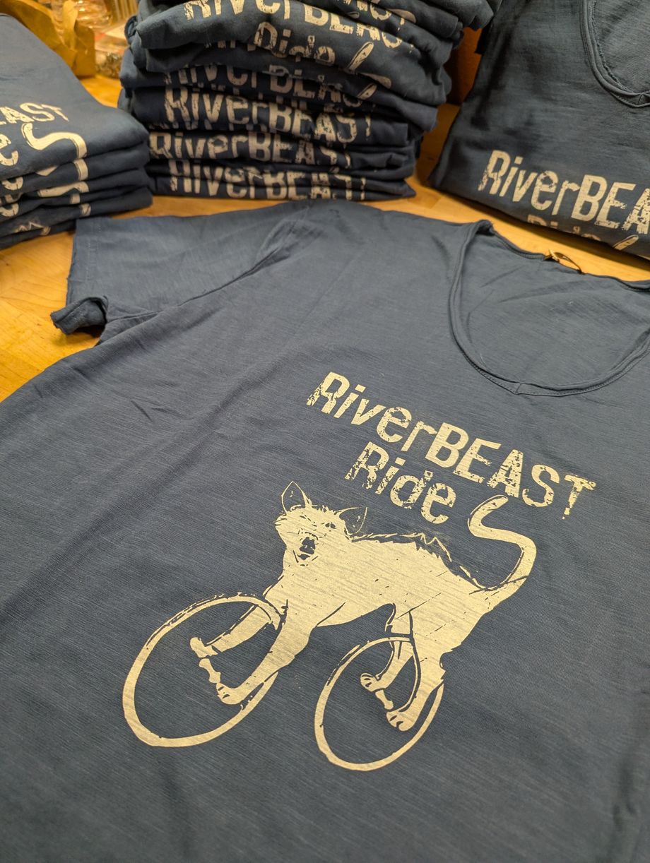 Stacks of slate blue RiverBEAST Ride shirts, one laid flat showing the distressed cat-on-a-bicycle print in cream ink